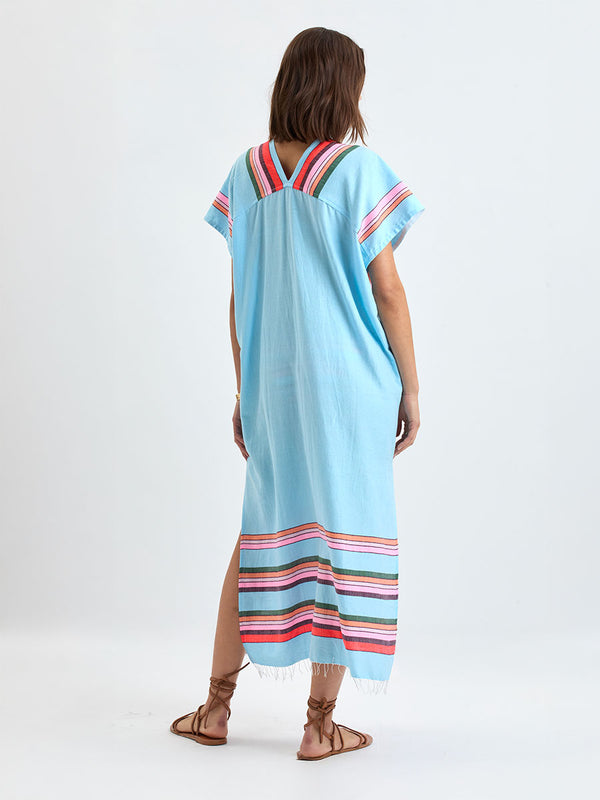 Back of a woman wearing lemlem GASIRA Caftan featuring  bold stripes in soft pink, tangerine, scarlet, forest green, and burgundy, set against a light aqua background.