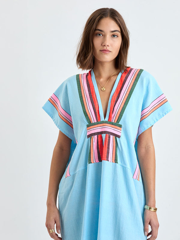 Close up on a woman wearing lemlem GASIRA Caftan featuring  bold stripes in soft pink, tangerine, scarlet, forest green, and burgundy, set against a light aqua background.