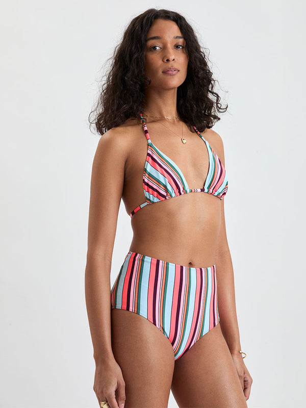 Woman wearing lemlem MALIA Triangle Top and ELSI High Waist Bottom featuring  bold stripes in soft pink, tangerine, scarlet, forest green, and burgundy, set against a light aqua background.