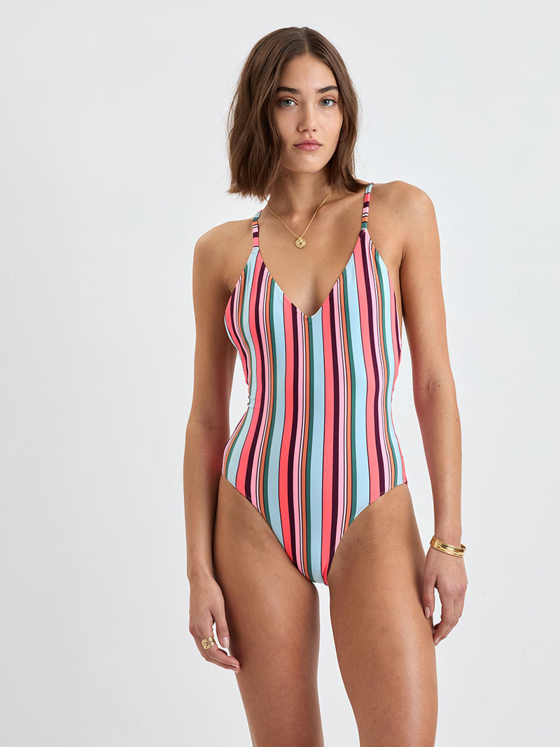 Woman wearing lemlem RETIRO One Piece featuring  bold stripes in soft pink, tangerine, scarlet, forest green, and burgundy, set against a light aqua background.