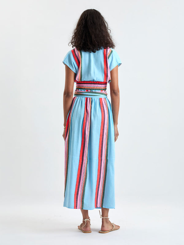 Back of a woman wearing lemlem ALIA Plunge Top and TOLA Skirt featuring color block stripe pattern in white on sky blue background