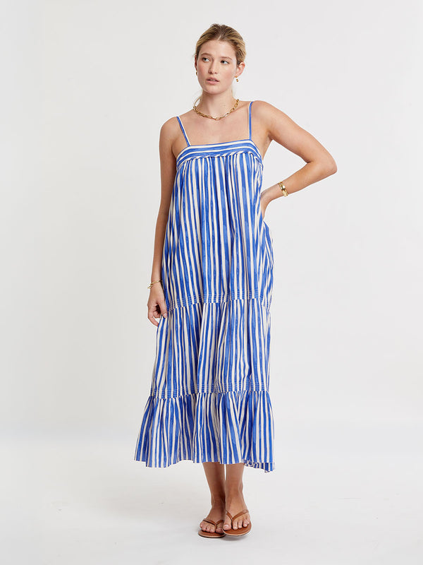 Woman standing wearing lemlem BEZA Dress featuring an exclusive in-house designed painted-effect stripe print in a vibrant, dazzling blue on a white background.