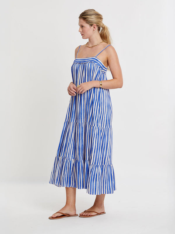 Woman standing wearing lemlem BEZA Dress featuring an exclusive in-house designed painted-effect stripe print in a vibrant, dazzling blue on a white background.