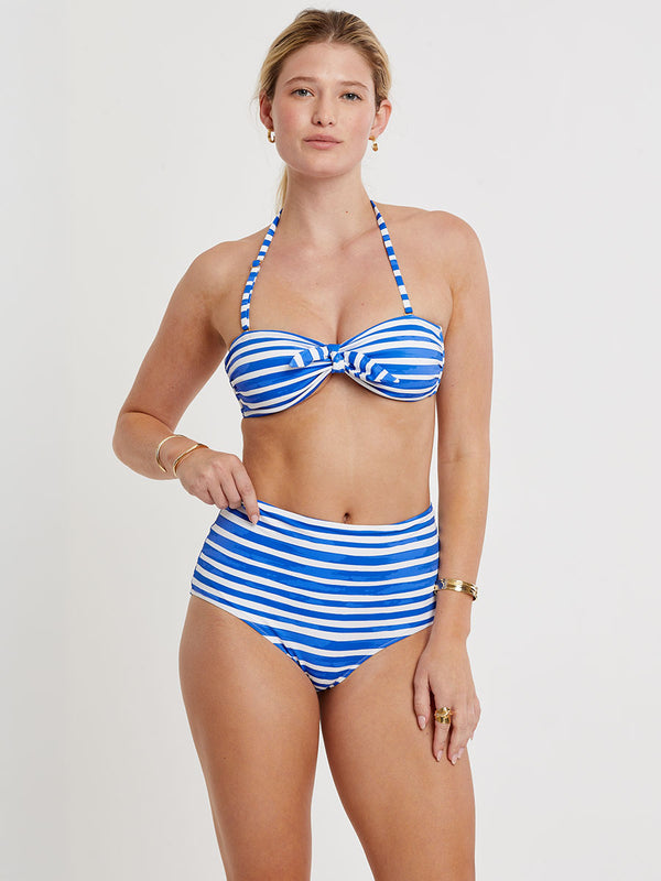 Woman standing wearing lemlem AVA Bandeau Top and ELSI High Waist Bottom featuring an exclusive in-house designed painted-effect stripe print in a vibrant, dazzling blue on a white background.