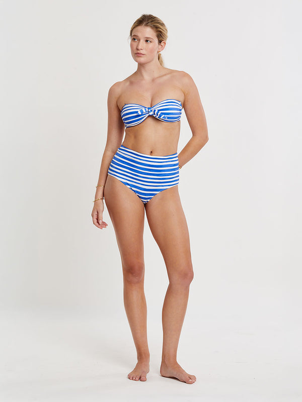 Woman standing wearing lemlem AVA Bandeau Top and ELSI High Waist Bottom featuring an exclusive in-house designed painted-effect stripe print in a vibrant, dazzling blue on a white background.