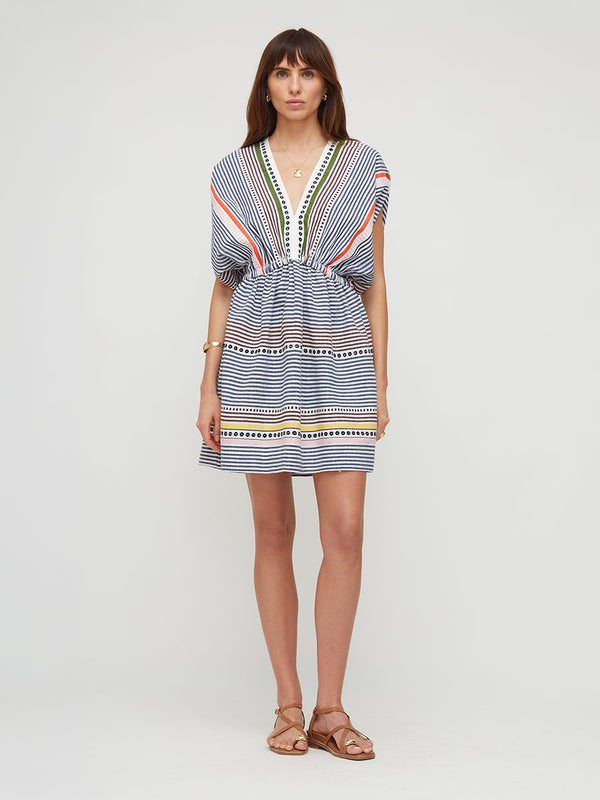A woman modeling a short, loose-fitting lemlem ALEM handwoven dress with a blue, white, and orange striped pattern, featuring a V-neckline with green and white trim, styled with brown strappy sandals against a light gray studio background.