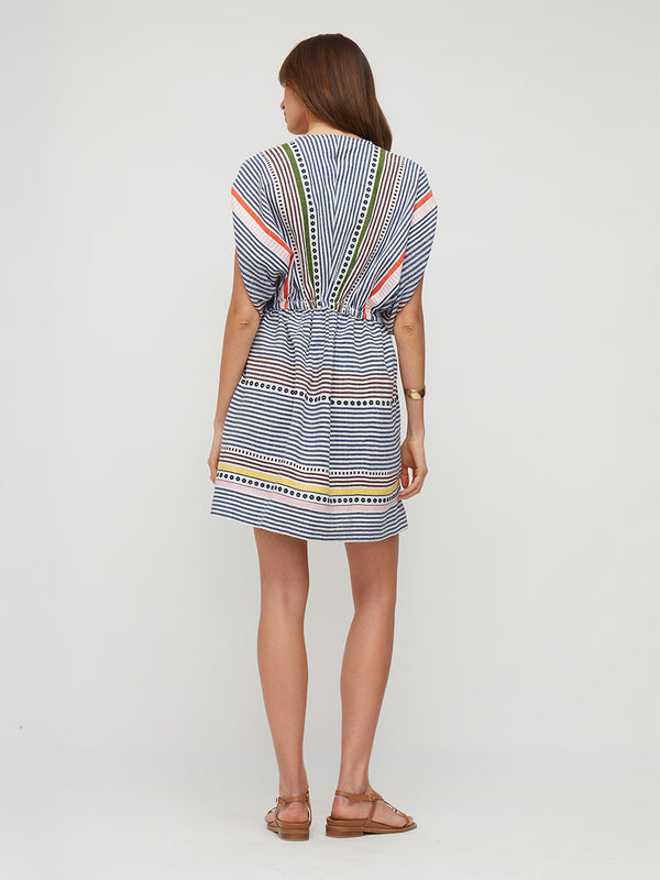 A woman modeling a short, loose-fitting lemlem ALEM handwoven dress with a blue, white, and orange striped pattern, featuring a V-neckline with green and white trim, styled with brown strappy sandals against a light gray studio background.