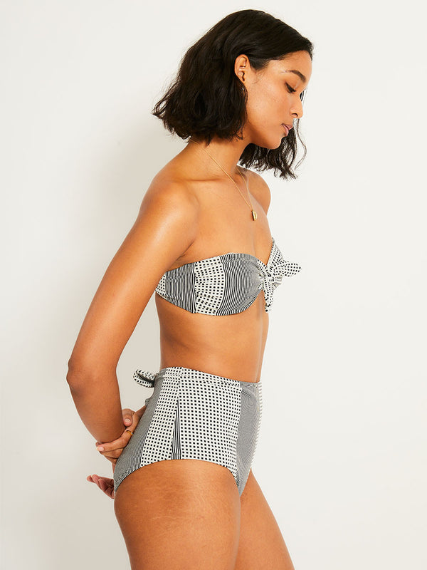 Side shot of a Woman Standing Wearing lemlem AVA Bandeau Top  featuring archive signature handwoven Semira patter in black and ivory colors and matching high waist bottom
