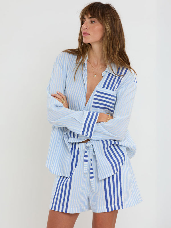 Woman Standing wearing lemlem  handwoven AISHA Shirt and SAFIA Shorts featuring  layered stripe pattern combining crisp deep-blue lines with softer, washed sky-blue stripes and MALIA Triangle Top.
