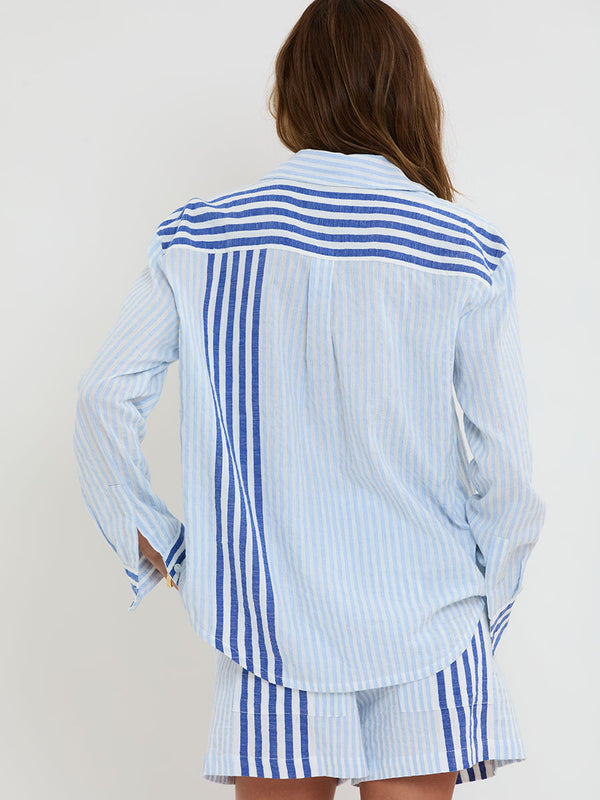 Woman Standing wearing lemlem  handwoven AISHA Shirt and SAFIA Shorts featuring  layered stripe pattern combining crisp deep-blue lines with softer, washed sky-blue stripes and MALIA Triangle Top.