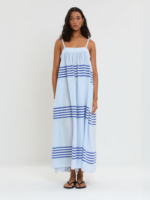 Woman Standing wearing lemlem  handwoven EDA Slip Dress featuring  layered stripe pattern combining crisp deep-blue lines with softer, washed sky-blue stripes and MALIA Triangle Top.