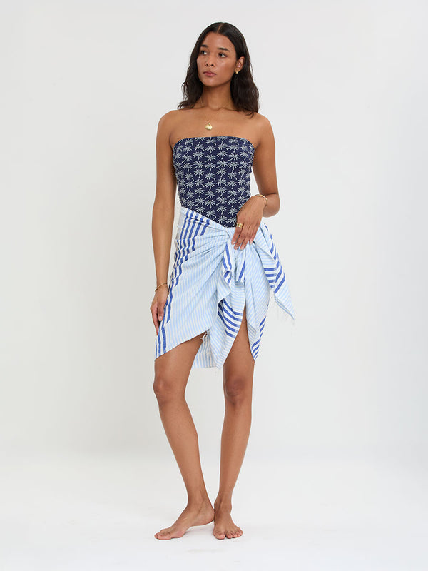 Woman Standing wearing lemlem handwoven LEMA Sarong featuring layered stripe pattern combining crisp deep-blue lines with softer, washed sky-blue stripes and DINHA One Piece swimsuit.