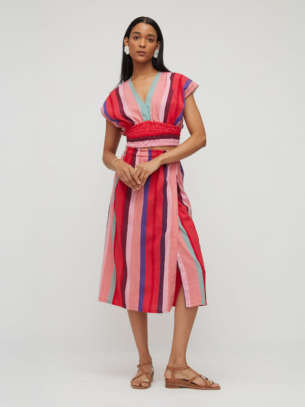 A woman modeling a short-sleeve midi dress with vertical pink, red, and light blue stripes, featuring a smocked waist and a split skirt, against a plain light gray studio background.