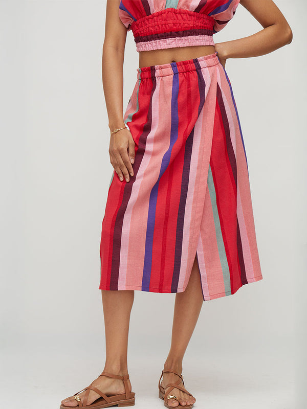 A model wearing a vibrant, multi-colored striped midi skirt with vertical bands of pink, red, purple, and teal, paired with brown leather sandals against a light gray studio background.
