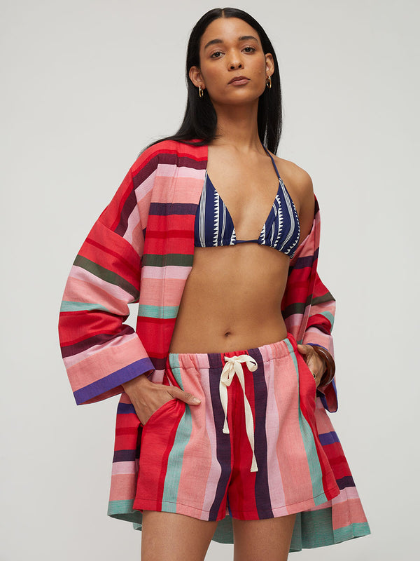 A model wearing a matching two-piece swimwear set featuring a pink, red, and teal striped bikini top and matching striped swim shorts, styled with a matching striped open-front cover-up against a light gray studio background.