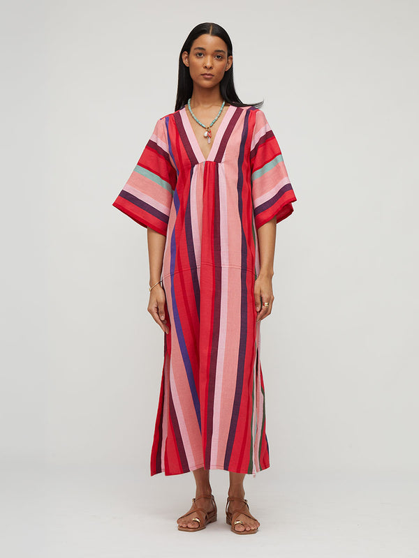 A woman modeling a loose, knee-length kaftan dress with vertical pink, red, and navy stripes, styled with a gold necklace and brown leather sandals against a light gray studio background.