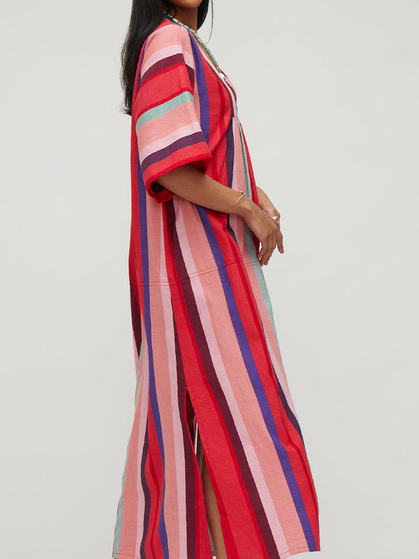 A woman wearing a loose, long kaftan dress with vertical multicolor stripes in red, pink, blue, and purple, shown from the side against a plain light gray studio background.