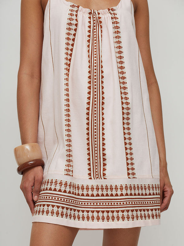 A woman modeling a sleeveless pink lemlem handwoven ZINA mini dress with brown crochet embroidery and fringe details, paired with brown leather sandals, against a light gray studio background