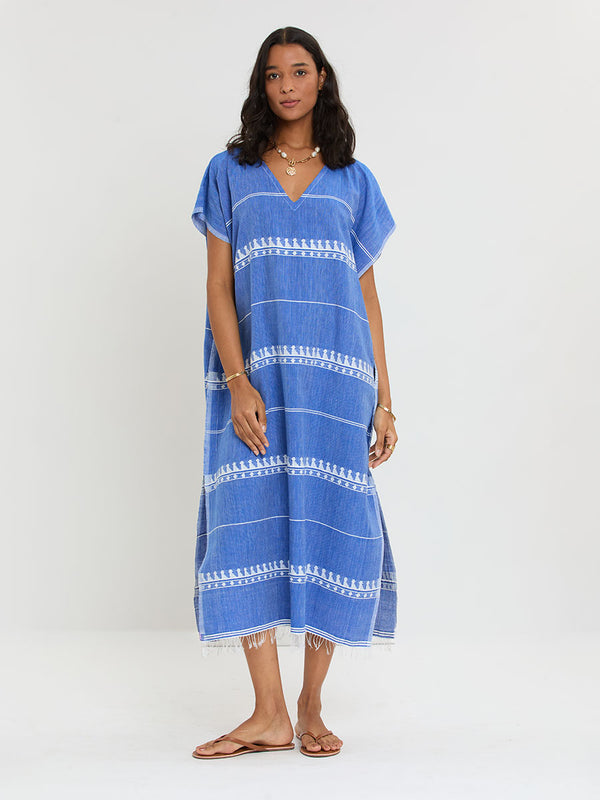 Woman Standing Wearing lemlem handwoven DALILA Caftan featuring an intricate weave of carefully placed tibebs in a crisp white color against a deep chambray blue background.