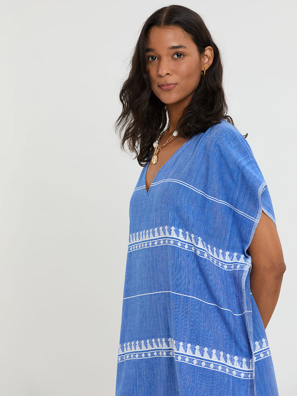 Woman Standing Wearing lemlem handwoven DALILA Caftan featuring an intricate weave of carefully placed tibebs in a crisp white color against a deep chambray blue background.