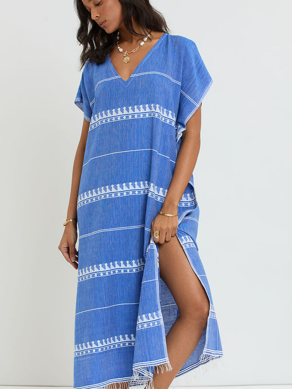 Woman Standing Wearing lemlem handwoven DALILA Caftan featuring an intricate weave of carefully placed tibebs in a crisp white color against a deep chambray blue background.