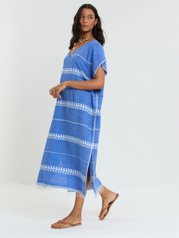 Woman Standing Wearing lemlem handwoven DALILA Caftan featuring an intricate weave of carefully placed tibebs in a crisp white color against a deep chambray blue background.