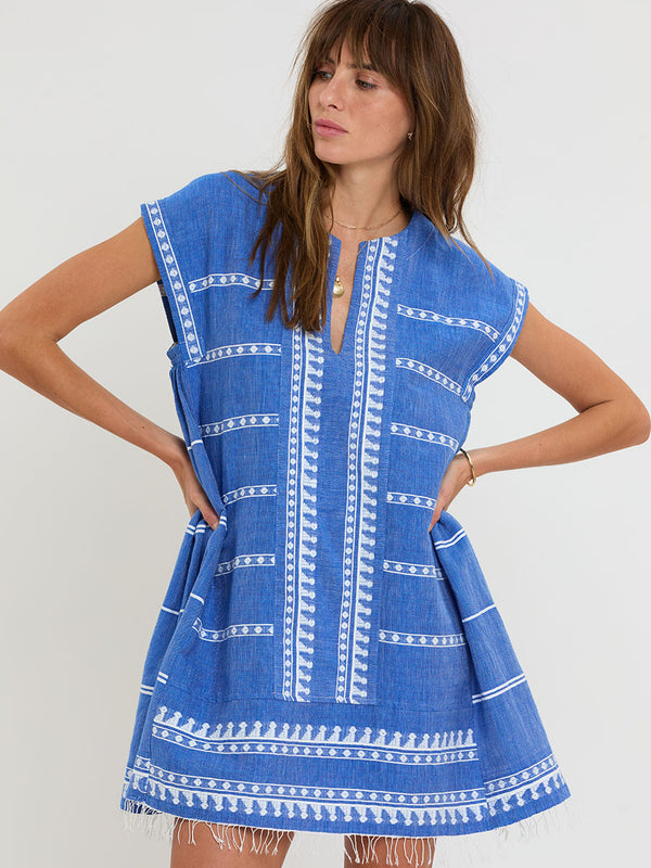 Woman Standing Wearing lemlem handwoven RAEY Column Dress featuring an intricate weave of carefully placed tibebs in a crisp white color against a deep chambray blue background.