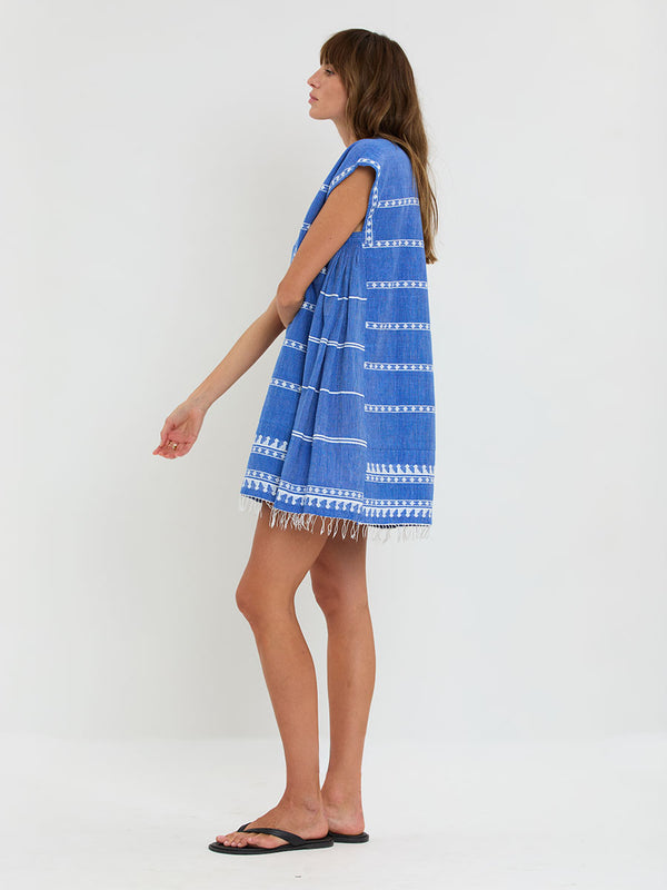 Woman Standing Wearing lemlem handwoven ELINA Caftan featuring an intricate weave of carefully placed tibebs in a crisp white color against a deep chambray blue background.