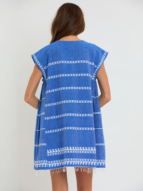 Woman Standing Wearing lemlem handwoven RAEY Column Dress featuring an intricate weave of carefully placed tibebs in a crisp white color against a deep chambray blue background.