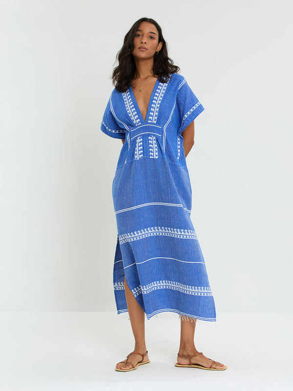Woman Standing Wearing lemlem handwoven GASIRA Caftan  featuring an intricate weave of carefully placed tibebs in a crisp white color against a deep chambray blue background 