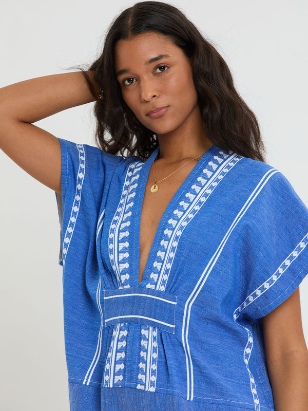 Woman Standing Wearing lemlem handwoven GASIRA Caftan  featuring an intricate weave of carefully placed tibebs in a crisp white color against a deep chambray blue background 