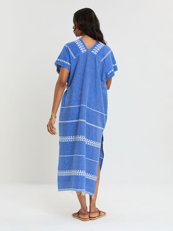Woman Standing Wearing lemlem handwoven GASIRA Caftan  featuring an intricate weave of carefully placed tibebs in a crisp white color against a deep chambray blue background 
