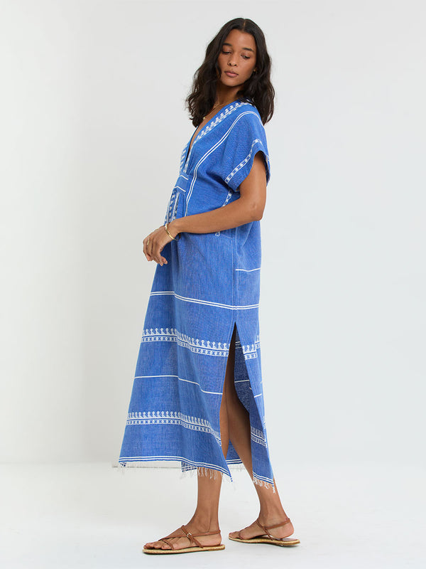Woman Standing Wearing lemlem handwoven GASIRA Caftan  featuring an intricate weave of carefully placed tibebs in a crisp white color against a deep chambray blue background 