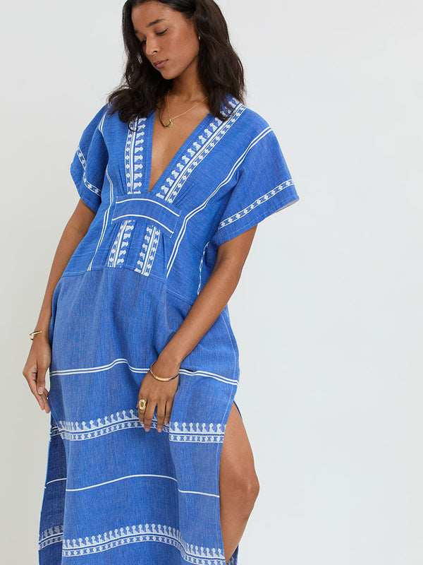 Woman Standing Wearing lemlem handwoven GASIRA Caftan  featuring an intricate weave of carefully placed tibebs in a crisp white color against a deep chambray blue background 