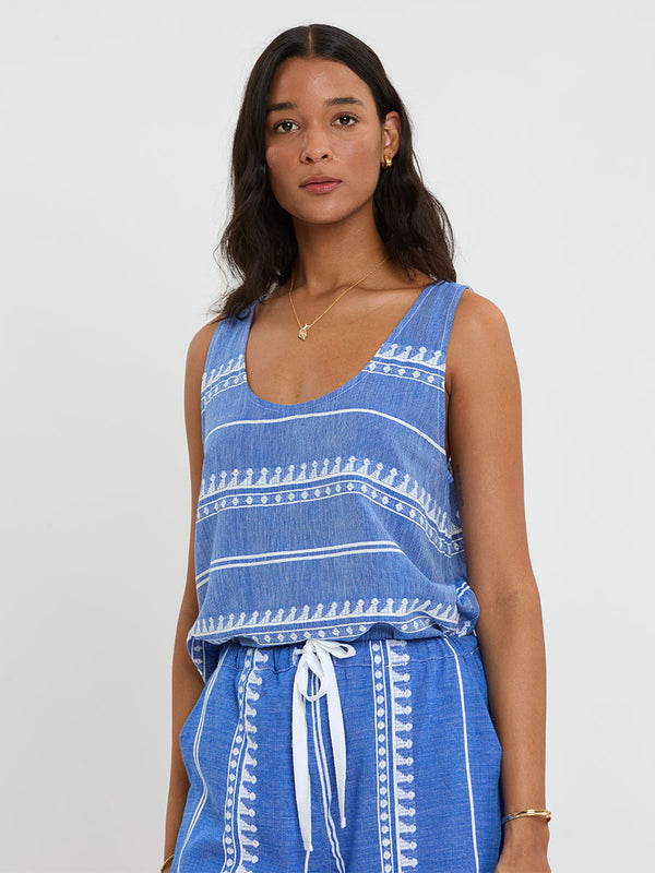 Woman Standing Wearing lemlem handwoven KATI Tank Top and DESTA Pants featuring an intricate weave of carefully placed tibebs in a crisp white color against a deep chambray blue background 