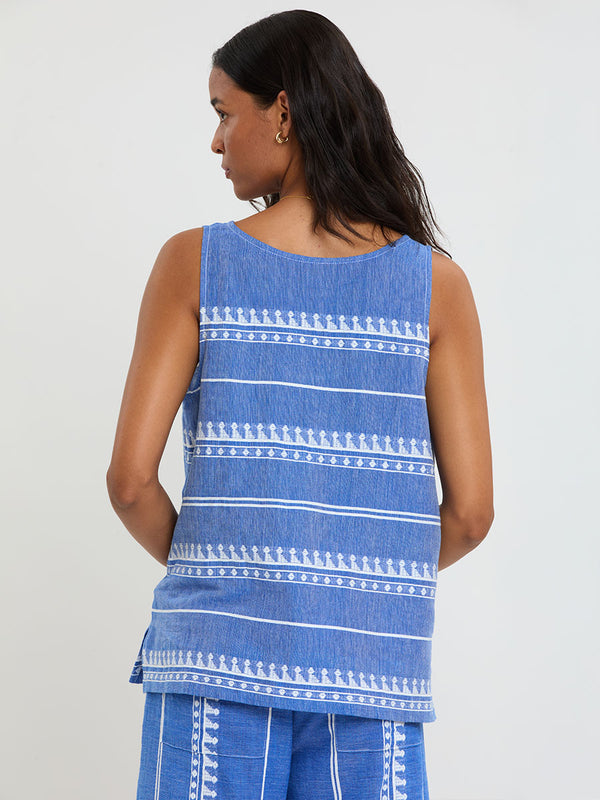 Back of a Woman Standing Wearing lemlem handwoven KATI Tank Top and DESTA Pants featuring an intricate weave of carefully placed tibebs in a crisp white color against a deep chambray blue background 