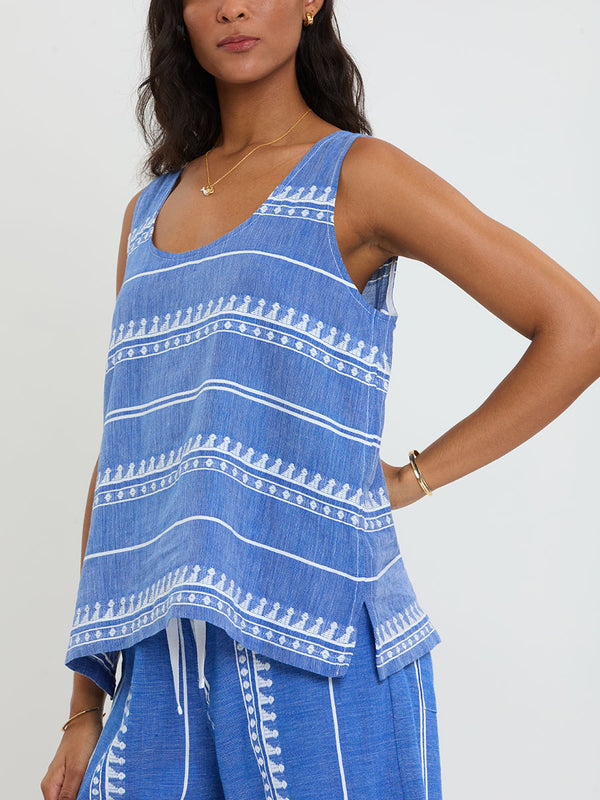 Woman Standing Wearing lemlem handwoven KATI Tank Top and DESTA Pants featuring an intricate weave of carefully placed tibebs in a crisp white color against a deep chambray blue background 