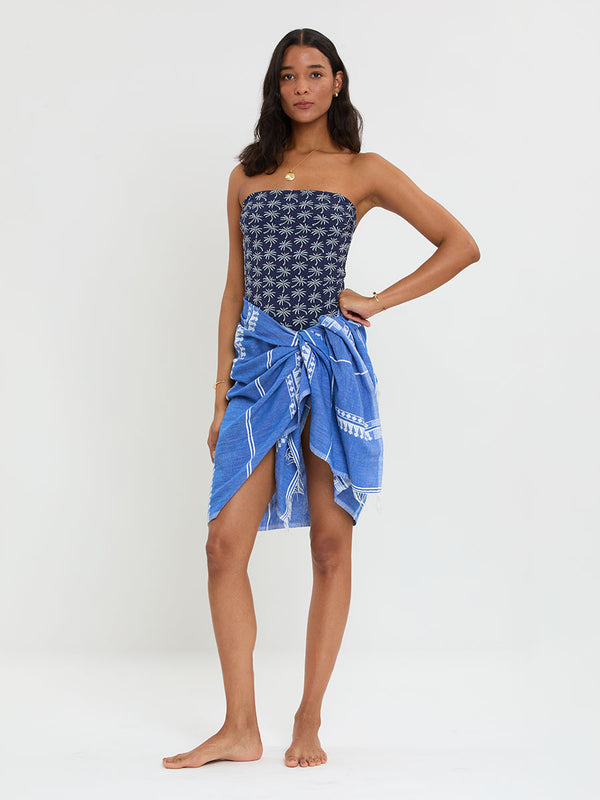 Woman Standing Wearing lemlem handwoven LEMA Sarong featuring an intricate weave of carefully placed tibebs in a crisp white color against a deep chambray blue background and lemlem strapless one piece 