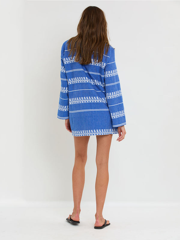 Back of a Woman Standing Wearing lemlem handwoven RAEY Column Dress featuring an intricate weave of carefully placed tibebs in a crisp white color against a deep chambray blue background.