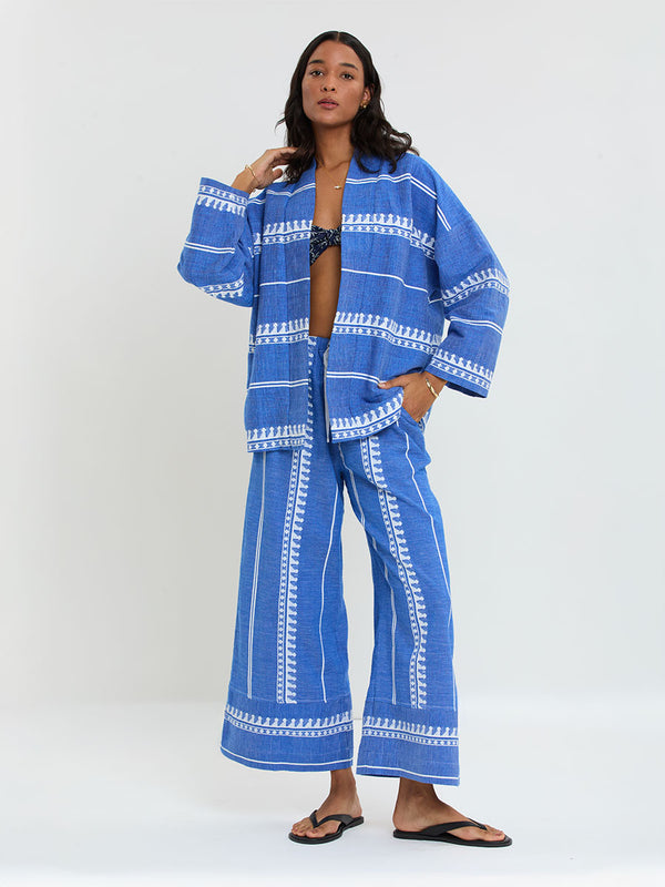 Woman Standing Wearing lemlem handwoven TERARA Jacket and DESTA Pants featuring an intricate weave of carefully placed tibebs in a crisp white color against a deep chambray blue background 