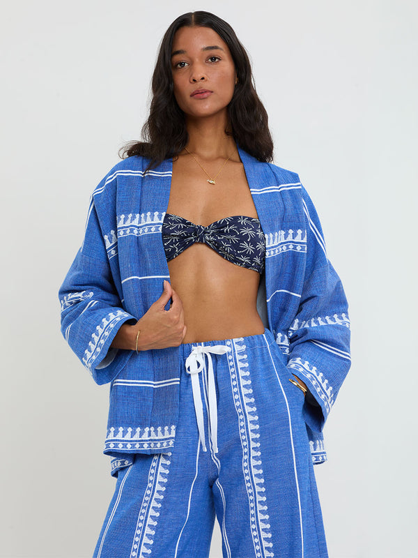 Woman Standing Wearing lemlem handwoven TERARA Jacket and DESTA Pants featuring an intricate weave of carefully placed tibebs in a crisp white color against a deep chambray blue background 