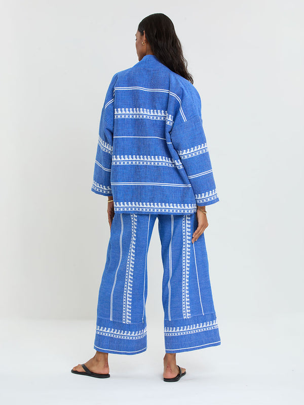 Woman Standing Wearing lemlem handwoven TERARA Jacket and DESTA Pants featuring an intricate weave of carefully placed tibebs in a crisp white color against a deep chambray blue background 