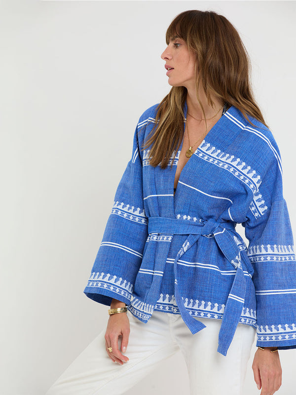 Woman Standing Wearing lemlem handwoven TERARA Jacket and DESTA Pants featuring an intricate weave of carefully placed tibebs in a crisp white color against a deep chambray blue background 