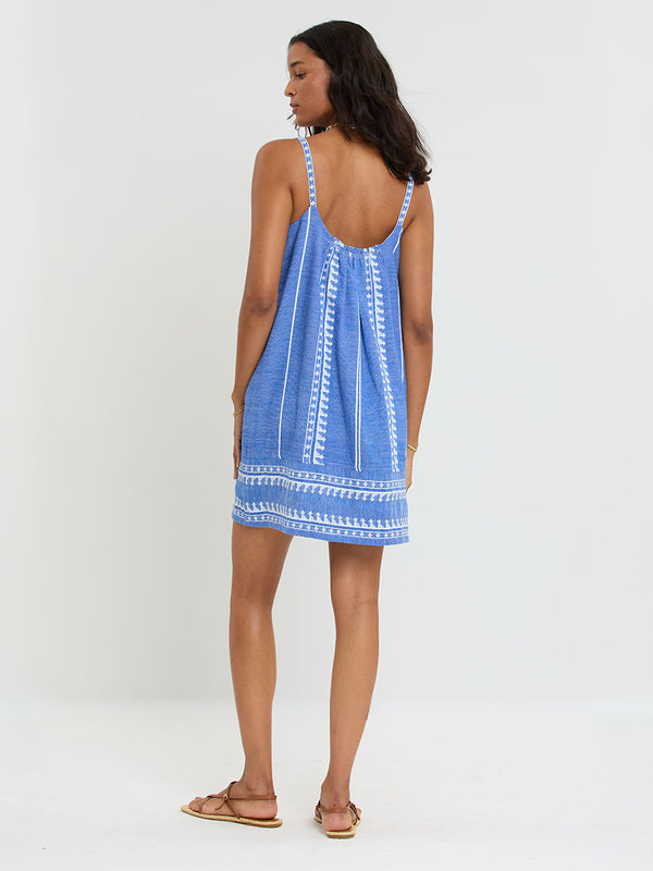 Woman Standing Wearing lemlem handwoven ZINA Dress featuring an intricate weave of carefully placed tibebs in a crisp white color against a deep chambray blue background.