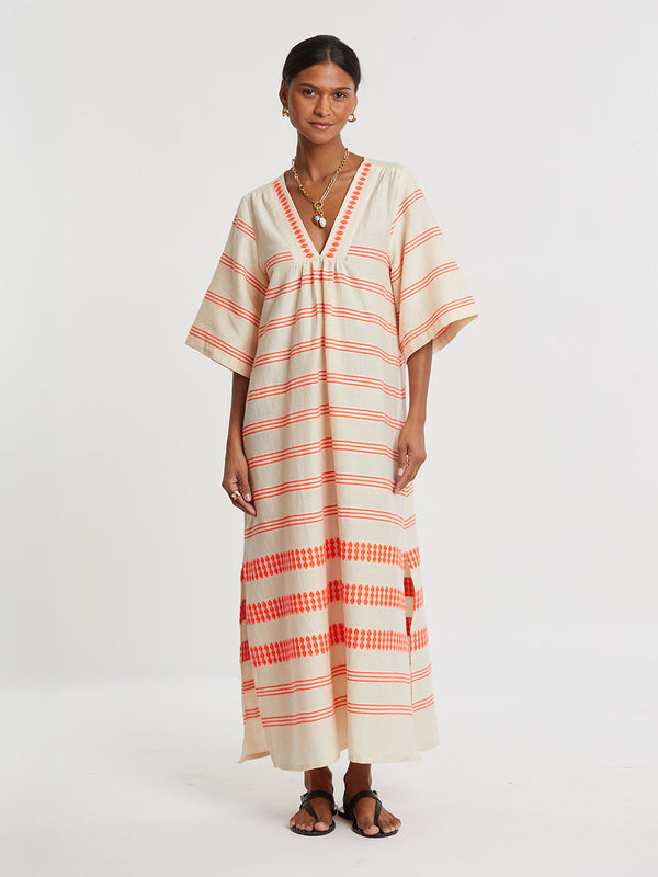 Woman Standing wearing lemlem handwoven EDNA Dress featuring intricate electric orange toned Tibeb pattern on a cream biscuit background.