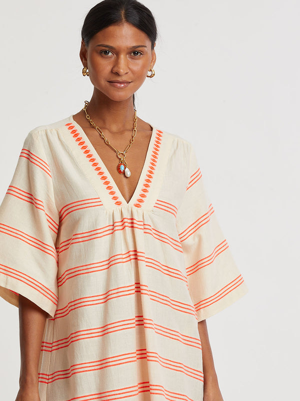 Woman Standing wearing lemlem handwoven EDNA Dress featuring intricate electric orange toned Tibeb pattern on a cream biscuit background.