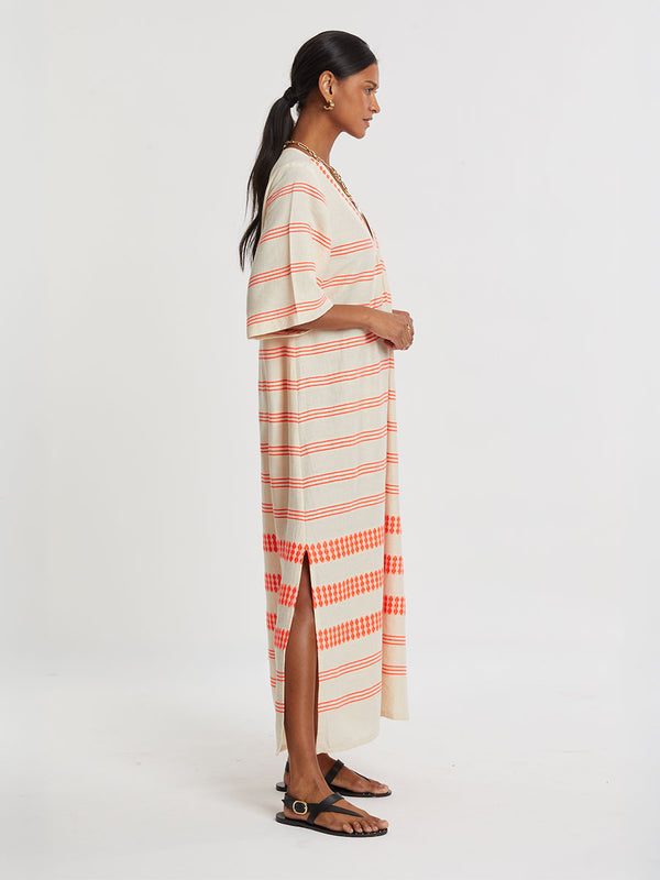 Woman Standing wearing lemlem handwoven EDNA Dress featuring intricate electric orange toned Tibeb pattern on a cream biscuit background.