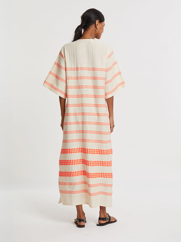 Woman Standing wearing lemlem handwoven EDNA Dress featuring intricate electric orange toned Tibeb pattern on a cream biscuit background.