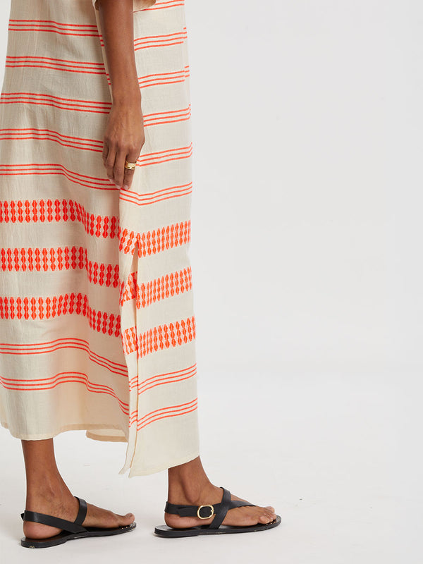 Woman Standing wearing lemlem handwoven EDNA Dress featuring intricate electric orange toned Tibeb pattern on a cream biscuit background.