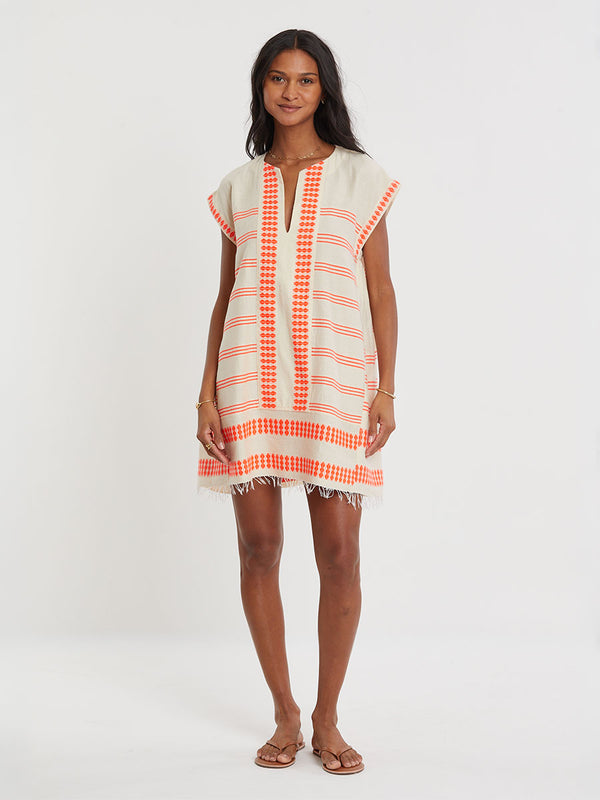 Woman Standing wearing lemlem handwoven ELINA Caftan featuring intricate electric orange toned Tibeb pattern on a cream biscuit background.
